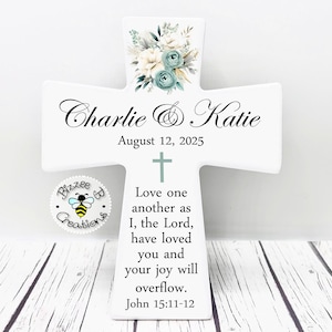 May include: A white ceramic cross featuring a floral arrangement in blue and cream tones at the top. It is personalized with the names Charlie & Katie, the date August 12, 2025, and the scripture verse John 15:11-12 in black lettering.