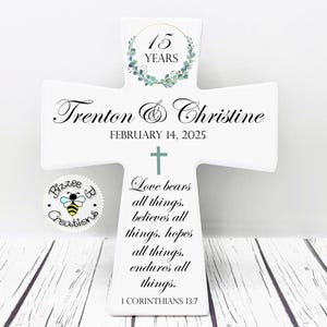 May include: White wooden cross with a green wreath and the text "15 Years" on the top. The cross also has the text "Trenton & Christine" and "February 14, 2025" on the front. The bottom of the cross has a Bible verse from 1 Corinthians 13:7.