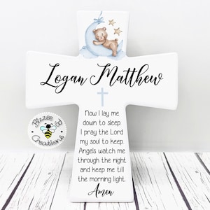 May include: White wooden cross with a blue and white illustration of a teddy bear sleeping on a crescent moon. The cross has the names "Logan Matthew" and the text "Now I lay me down to sleep. I pray the Lord my soul to keep. Angels watch me through the night and keep me till the morning light. Amen."