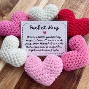 May include: A card with the text "Pocket Hug" and the message "Here's a little pocket hug. Keep it close all warm and snug. Even though I can't be there, you can squeeze this tight and know I care."  The card is surrounded by six small crocheted hearts in various shades of pink, red, and white.