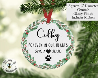 May include: White ceramic Christmas ornament with a green and red holly wreath, the name "Colby", the text "Forever in our hearts", the years "2002" and "2020", a heart, and a paw print. The ornament is 3 inches in diameter and includes a red ribbon.