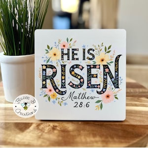 May include: White wooden sign with floral design and the text "He is Risen" in black lettering. The sign also includes the text "Matthew 28:6" in black lettering.
