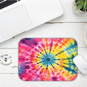 May include: A colorful tie-dye mouse pad with a circular pattern in shades of pink, yellow, blue, and green. The mouse pad is on a white wooden surface.
