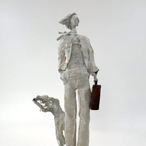May include: A white textured sculpture of a standing figure wearing a jacket and trousers, holding a small brown rectangular case, accompanied by a smaller white textured figure, all mounted on a square light brown wooden base.