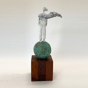May include: A white wire-sculpted figure stands atop a textured, spherical green patina base, which is mounted on a solid, dark brown rectangular wooden block. The figure has an elongated form with one arm extended horizontally to the side.