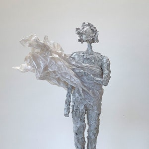 May include: A textured, gray sculpture of a figure holding a flowing, white fabric. The figure stands on a small, square wooden base. The sculpture has a rough, almost weathered appearance, with visible texture and detail.