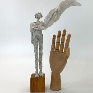 May include: A white textured sculpture of a slender figure standing on a square wooden base, draped in a thin, flowing white fabric. Beside it stands a light-colored wooden articulated hand model with visible joints, fingers, and a thumb.