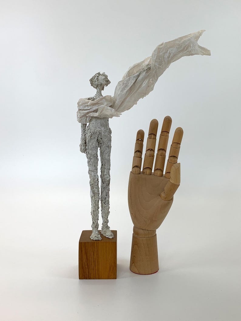 May include: A white textured sculpture of a slender figure standing on a square wooden base, draped in a thin, flowing white fabric. Beside it stands a light-colored wooden articulated hand model with visible joints, fingers, and a thumb.