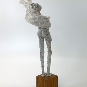 May include: A sculpture of a slender, textured grey figure standing on a solid, square wooden base. The figure has a rough, uneven surface and is draped in a thin, crinkled, off-white material that extends outward from the upper body area.