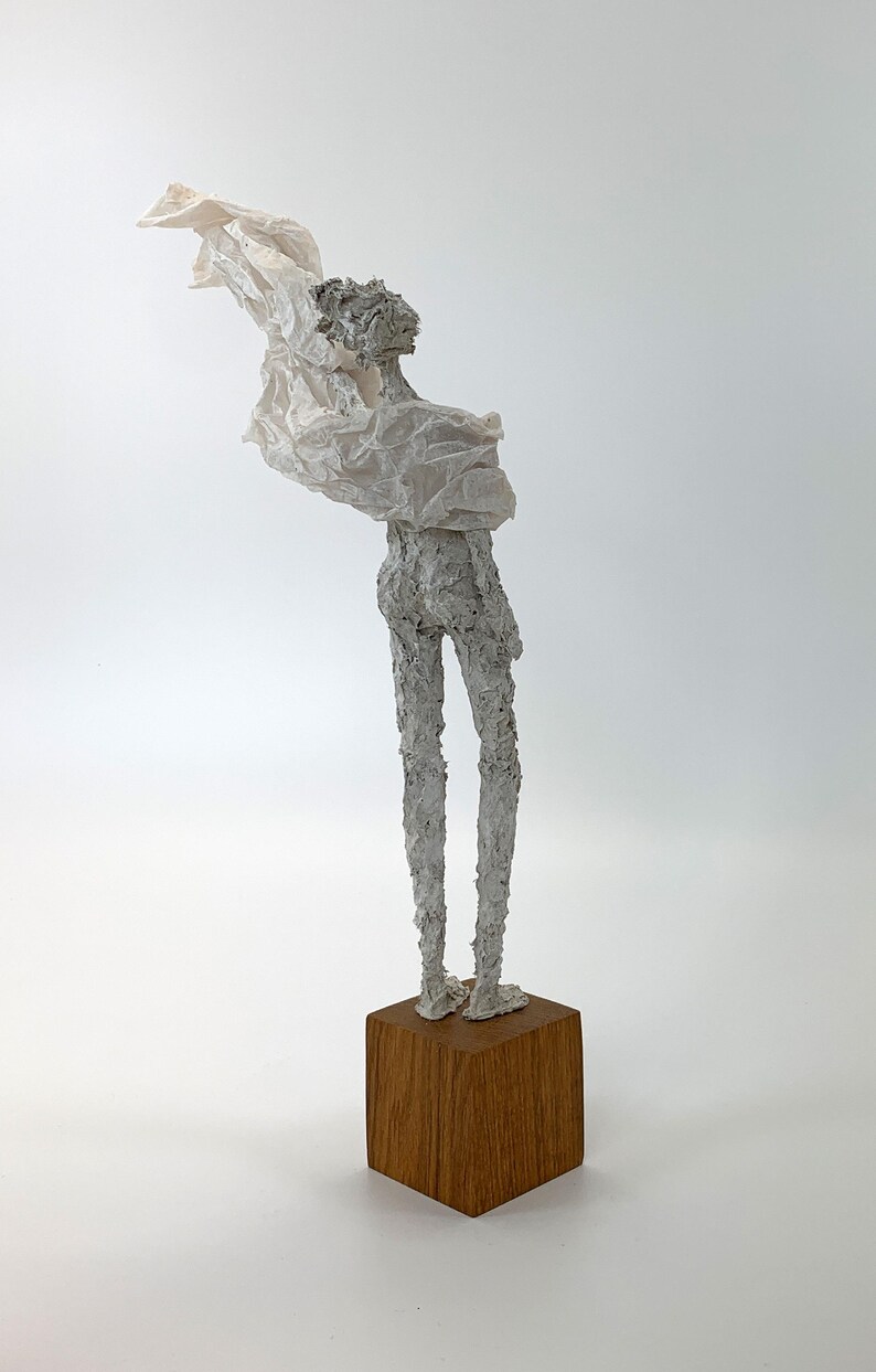 May include: A sculpture of a slender, textured grey figure standing on a solid, square wooden base. The figure has a rough, uneven surface and is draped in a thin, crinkled, off-white material that extends outward from the upper body area.