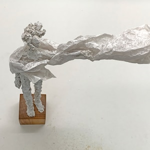 May include: A white textured sculpture of a figure standing on a square wooden base. The figure has a rough, crinkled surface and is draped in a long, flowing, translucent white material that extends horizontally to the right side of the frame.