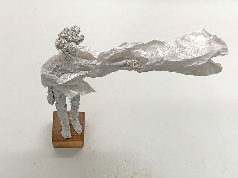 May include: A white textured sculpture of a figure standing on a square wooden base. The figure has a rough, crinkled surface and is draped in a long, flowing, translucent white material that extends horizontally to the right side of the frame.