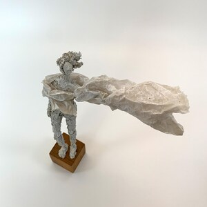 May include: A textured white sculpture of a figure standing on a square wooden base. The figure has a rough, granular surface and is draped in a large, translucent, crinkled white material that extends outward to one side like a flowing cape.