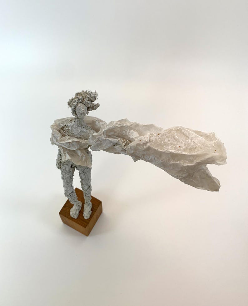 May include: A textured white sculpture of a figure standing on a square wooden base. The figure has a rough, granular surface and is draped in a large, translucent, crinkled white material that extends outward to one side like a flowing cape.