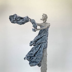 May include: A white sculpture of a person with a flowing blue fabric draped over their body. The sculpture is standing on a white pedestal.