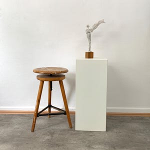 Eine monochrome, aufrechte Skulptur mit einer wehenden Stola auf einem massiven Eichensockel steht auf einem weißen Galerie-Sockel, daneben befindet sich ein minimalistischer Holzhocker vor einer hellen Wand.