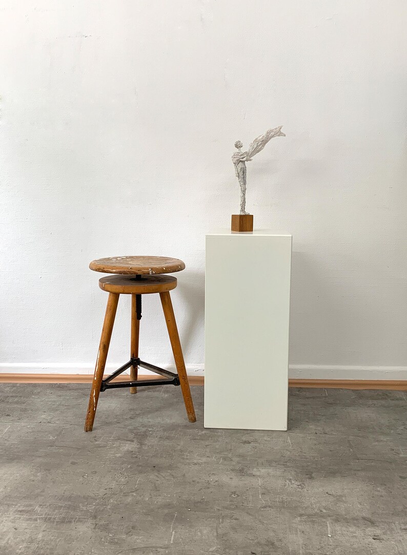 Eine monochrome, aufrechte Skulptur mit einer wehenden Stola auf einem massiven Eichensockel steht auf einem weißen Galerie-Sockel, daneben befindet sich ein minimalistischer Holzhocker vor einer hellen Wand.