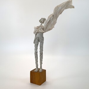 May include: A tall, slender sculpture of a figure with a textured, off-white surface standing on a square wooden base. The figure has a thin, elongated form and is draped in a light, translucent, off-white fabric that extends outward.
