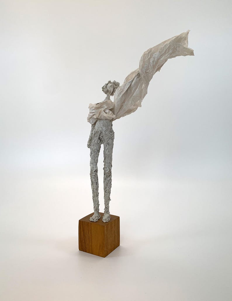 May include: A tall, slender sculpture of a figure with a textured, off-white surface standing on a square wooden base. The figure has a thin, elongated form and is draped in a light, translucent, off-white fabric that extends outward.