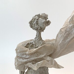 May include: A textured, light gray sculpture of a figure with a rough, organic surface. The figure has a rounded head and is draped in a translucent, off-white, crinkled material that extends outward from the shoulders, creating a flowing effect.