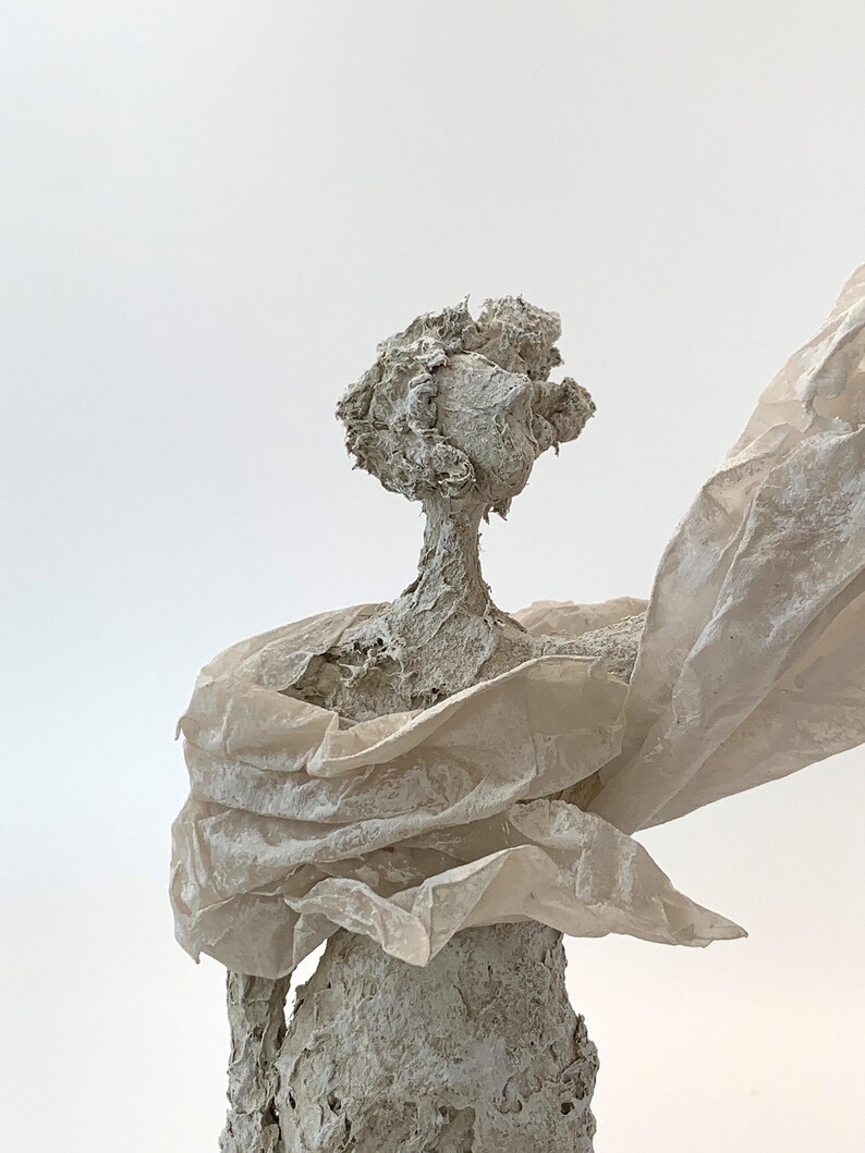 May include: A textured, light gray sculpture of a figure with a rough, organic surface. The figure has a rounded head and is draped in a translucent, off-white, crinkled material that extends outward from the shoulders, creating a flowing effect.