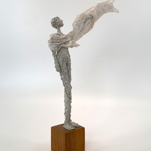 May include: A tall, slender sculpture of a figure with a textured, light gray surface standing on a square wooden base. The figure is draped in a thin, translucent, off-white fabric that extends outward from the shoulder, creating a sense of motion.