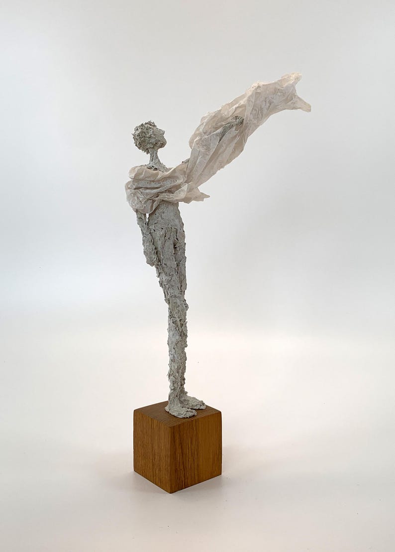 May include: A tall, slender sculpture of a figure with a textured, light gray surface standing on a square wooden base. The figure is draped in a thin, translucent, off-white fabric that extends outward from the shoulder, creating a sense of motion.