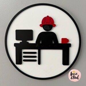 Könnte beinhalten: Ein rundes Wandschild mit weißem Hintergrund und schwarzem Rahmen zeigt die schwarze Silhouette einer Person mit rotem Feuerwehrhelm, die an einem Schreibtisch mit Computerbildschirm und roter Tasse sitzt. Der Text Annie & Paul steht unten rechts.
