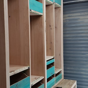 May include: A wooden cabinet with teal drawers and shelves. The cabinet is unfinished and has a light wood finish. The cabinet is being built in a workshop.