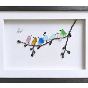 Op de afbeelding: Ingelijst kunstwerk met een tak met zeven kleurrijke glazen vogels die erop zitten. De vogels zijn in verschillende kleuren, waaronder roze, geel, blauw en groen. Er is ook een vlinder afgebeeld. Het kunstwerk is ingelijst in een zwart frame met een witte passe-partout.