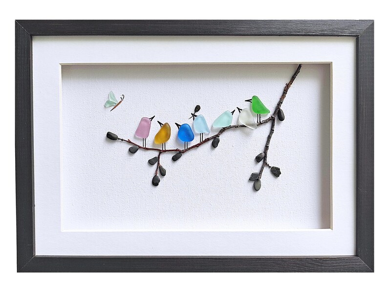 Op de afbeelding: Ingelijst kunstwerk met een tak met zeven kleurrijke glazen vogels die erop zitten. De vogels zijn in verschillende kleuren, waaronder roze, geel, blauw en groen. Er is ook een vlinder afgebeeld. Het kunstwerk is ingelijst in een zwart frame met een witte passe-partout.