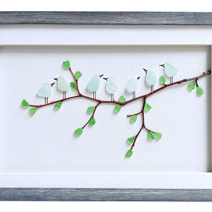 May include: A framed artwork featuring a branch with eight light blue glass birds perched on it. The branch has green glass leaves. The frame is gray and the background is white.