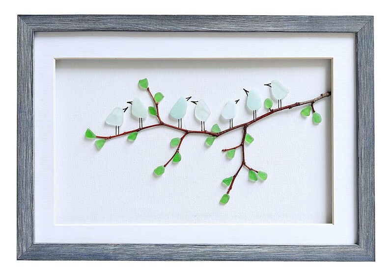 May include: A framed artwork featuring a branch with eight light blue glass birds perched on it. The branch has green glass leaves. The frame is gray and the background is white.