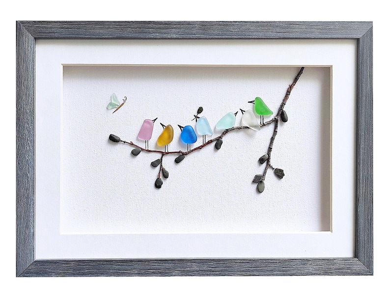 Op de afbeelding: Ingelijst kunstwerk met een tak met kleurrijke glazen vogels. De vogels zijn roze, geel, blauw, lichtblauw, groen en wit. Een lichtgroene vlinder staat linksboven. De lijst is grijs.