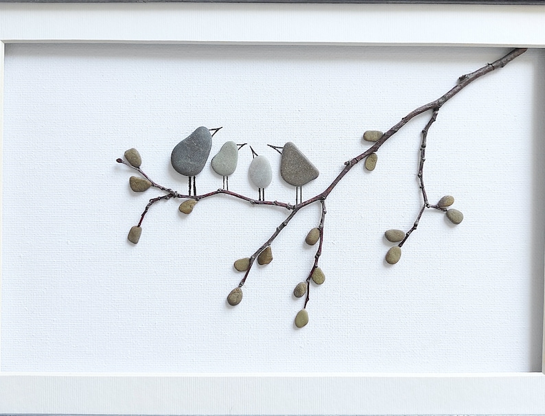 May include: A framed artwork featuring a twig with small brown pebbles arranged to look like leaves and gray pebbles arranged to look like birds perched on the twig.