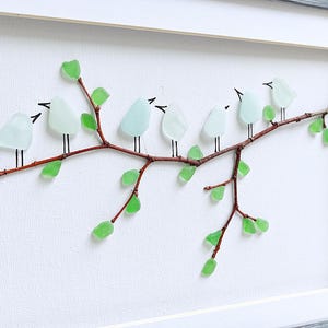 May include: A framed artwork featuring a branch with light blue glass birds perched on it. The branch has green glass leaves. The artwork is set against a white background and framed in a gray wooden frame.