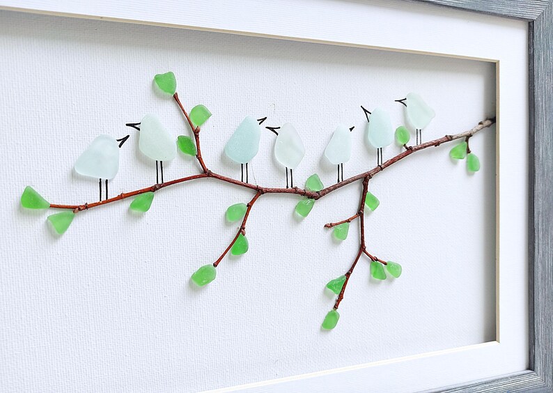 May include: A framed artwork featuring a branch with light blue glass birds perched on it. The branch has green glass leaves. The artwork is set against a white background and framed in a gray wooden frame.