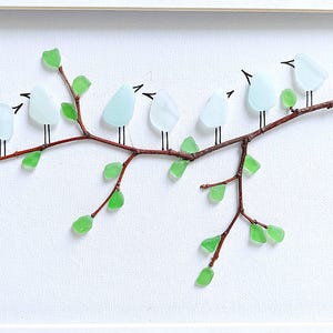 May include: A framed artwork featuring a branch with glass birds and leaves. The birds are light blue, the leaves are green, and the branch is brown. The birds are perched on the branch, and the leaves are scattered around it. The background is white.