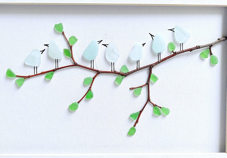 May include: A framed artwork featuring a branch with glass birds and leaves. The birds are light blue, the leaves are green, and the branch is brown. The birds are perched on the branch, and the leaves are scattered around it. The background is white.