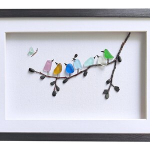 Op de afbeelding: Een ingelijst kunstwerk met een tak met zeven kleurrijke glazen vogels. De vogels zijn in verschillende kleuren, waaronder roze, geel, blauw en groen. Een kleine glazen vlinder bevindt zich in de linkerbovenhoek. Het kunstwerk zit in een zwarte lijst.