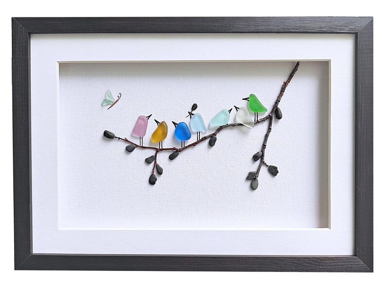 Op de afbeelding: Een ingelijst kunstwerk met een tak met zeven kleurrijke glazen vogels. De vogels zijn in verschillende kleuren, waaronder roze, geel, blauw en groen. Een kleine glazen vlinder bevindt zich in de linkerbovenhoek. Het kunstwerk zit in een zwarte lijst.