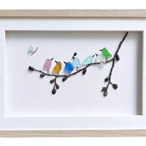 Op de afbeelding: Ingelijst kunstwerk met een tak met kleurrijke glazen vogels en een vlinder. De vogels zijn in tinten roze, geel, blauw, lichtblauw, wit en groen. De lijst is van licht hout en de achtergrond is wit.