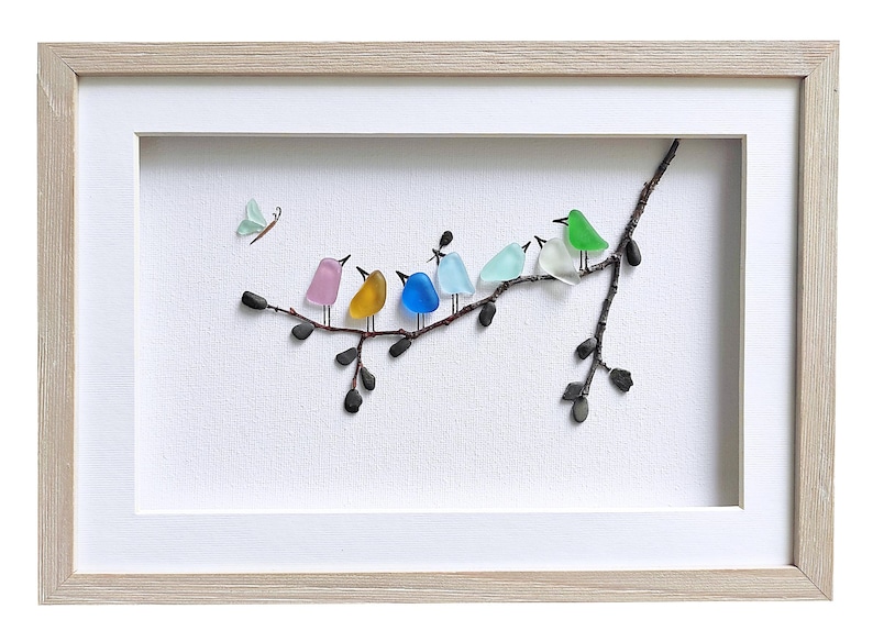 Op de afbeelding: Ingelijst kunstwerk met een tak met kleurrijke glazen vogels en een vlinder. De vogels zijn in tinten roze, geel, blauw, lichtblauw, wit en groen. De lijst is van licht hout en de achtergrond is wit.