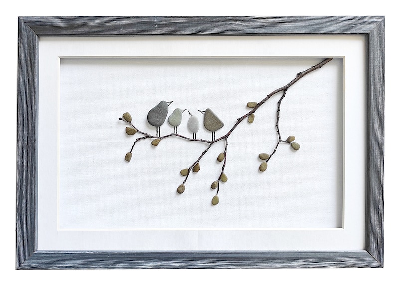 May include: A framed artwork featuring a pebble art design of four birds perched on a twig with leaves. The birds are gray and brown, and the leaves are green. The artwork is framed in a gray wood frame.