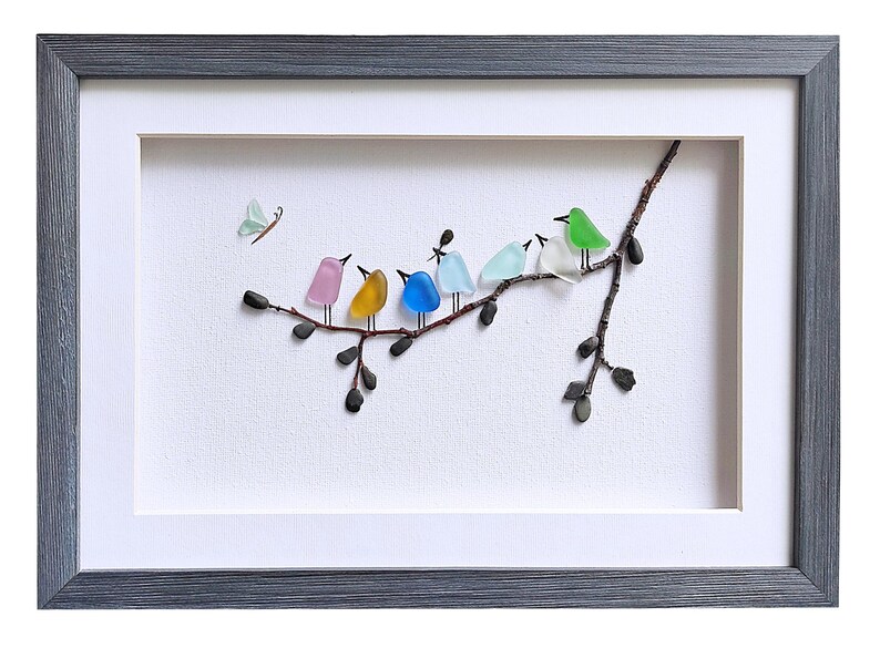 Op de afbeelding: Ingelijst kunstwerk met een tak met kleurrijke glazen vogels en een vlinder. De vogels zijn in roze, geel, blauw, lichtblauw, groen en wit. De lijst is grijs.