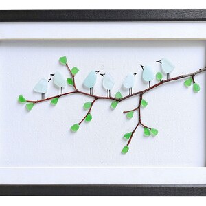 May include: Framed artwork featuring a branch with seven light blue bird figures made of glass. The birds are perched on a brown branch with green glass leaves. The artwork is set against a white background and framed in black.