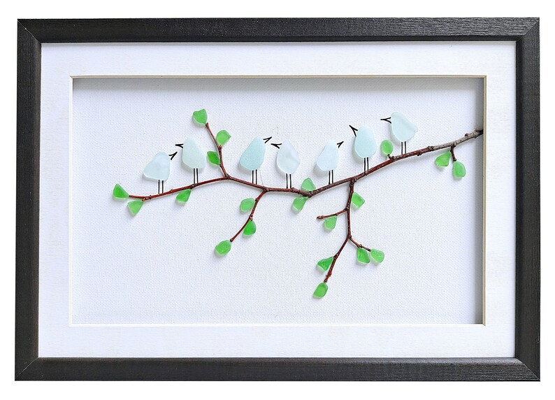 May include: Framed artwork featuring a branch with seven light blue bird figures made of glass. The birds are perched on a brown branch with green glass leaves. The artwork is set against a white background and framed in black.