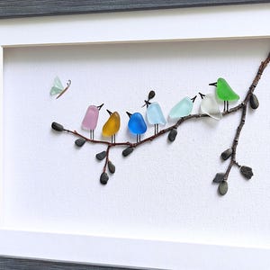 Op de afbeelding: Een ingelijst kunstwerk met een tak waarop kleurrijke glazen vogels zitten. De vogels hebben verschillende kleuren, waaronder roze, geel, blauw en groen. Er is ook een kleine vlinder aanwezig. De lijst is grijs en de achtergrond is wit.