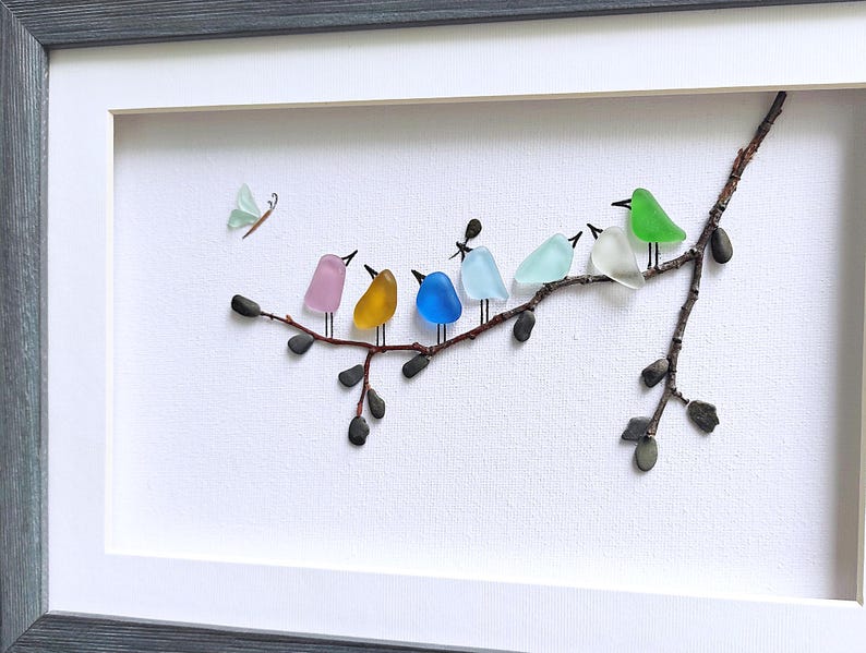 Op de afbeelding: Een ingelijst kunstwerk met een tak waarop kleurrijke glazen vogels zitten. De vogels hebben verschillende kleuren, waaronder roze, geel, blauw en groen. Er is ook een kleine vlinder aanwezig. De lijst is grijs en de achtergrond is wit.