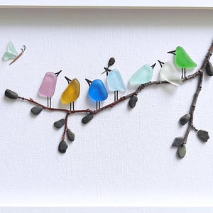 Op de afbeelding: Een ingelijst kunstwerk met een tak waarop kleurrijke glazen vogels zitten. De vogels zijn in roze, geel, blauw, lichtblauw, groen en wit. Er is ook een glazen vlinder aanwezig. Het kunstwerk staat tegen een witte achtergrond.
